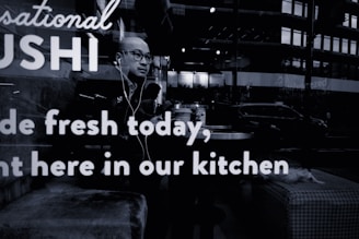 A person wearing glasses and earphones is seated at a table inside a sushi restaurant. The person appears to be looking outside through the window. Reflections from the street outside, including a few vehicles and buildings, are visible on the glass. The words 'Sushi made fresh today, right here in our kitchen' are prominently displayed on the window glass.