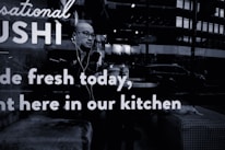 A person wearing glasses and earphones is seated at a table inside a sushi restaurant. The person appears to be looking outside through the window. Reflections from the street outside, including a few vehicles and buildings, are visible on the glass. The words 'Sushi made fresh today, right here in our kitchen' are prominently displayed on the window glass.