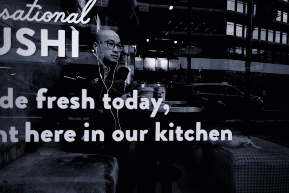 A person wearing glasses and earphones is seated at a table inside a sushi restaurant. The person appears to be looking outside through the window. Reflections from the street outside, including a few vehicles and buildings, are visible on the glass. The words 'Sushi made fresh today, right here in our kitchen' are prominently displayed on the window glass.