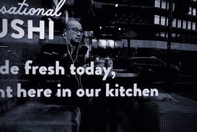 A person wearing glasses and earphones is seated at a table inside a sushi restaurant. The person appears to be looking outside through the window. Reflections from the street outside, including a few vehicles and buildings, are visible on the glass. The words 'Sushi made fresh today, right here in our kitchen' are prominently displayed on the window glass.