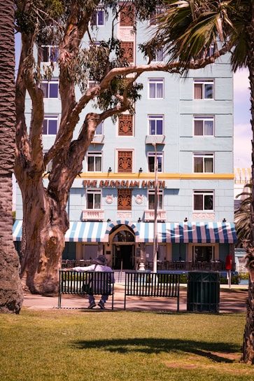 A building with a light blue facade adorned with decorative elements and several windows stands prominently. The awning is striped in blue and white, with a sign that reads 'The Georgian Hotel'. In front, a large tree with sprawling branches partially obscures the view. A few palm trees are visible, and a person sits alone on a bench, dressed in dark clothing with a wide-brimmed hat. The foreground features well-maintained green grass.