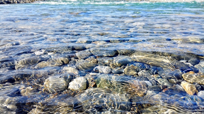 Clear water reveals smooth stones beneath, with gentle ripples capturing light reflections. The water appears crisp and blue at the top, transitioning to revealing stones of various grays and browns beneath.