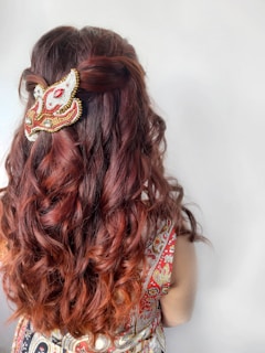 A person with long wavy brown hair viewed from the back, adorned with a decorative beaded hair accessory. The attire features a colorful and intricate pattern.