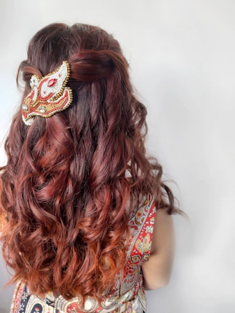 A person with long wavy brown hair viewed from the back, adorned with a decorative beaded hair accessory. The attire features a colorful and intricate pattern.