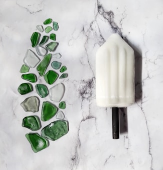 A white ice cream popsicle with a black stick lies on a marble surface next to various pieces of green and clear sea glass arranged in a gallery-like display.