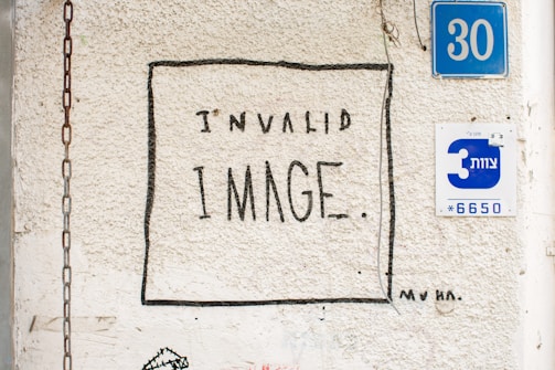 A textured wall with a hand-drawn box around the text 'INVALID IMAGE.' Nearby, there are two blue and white signs with numbers and Hebrew text, as well as a chain hanging on the wall.
