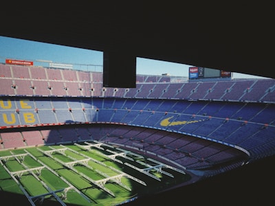 An empty, expansive football stadium with tiered seating adorned in blue and maroon colors. Green artificial turf with white structures covers the field, and advertising boards are visible on the stadium's upper sections.