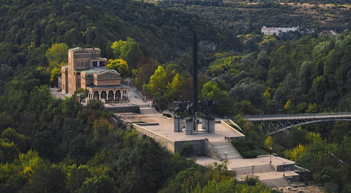 A historic building with a large, rectangular structure and arched openings sits amidst a dense forested hillside. In front, a prominent monument with equestrian sculptures and an obelisk-like feature is positioned on a wide, stepped platform. A bridge extends from the monument, crossing over a valley of lush greenery.