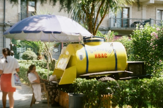A yellow tank labeled with Cyrillic letters and the word 'KBAC' is positioned on a cart. It is sheltered by a large blue and white beach umbrella. There are two women in the scene; one is standing and facing away from the camera, while the other is seated under the umbrella. The setting appears to be a sunny, outdoor area with lush greenery and an urban backdrop.