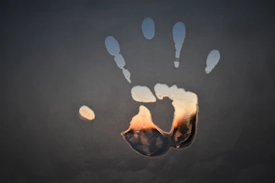 A handprint is imprinted on a foggy surface, with light seeping through the print, creating a colorful gradient effect. The background is dark, contrasting with the vibrant hues within the handprint.
