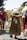 An elderly woman with white hair is walking uphill on a cobblestone path, holding a small bag containing red flowers behind her back. She is wearing a brown patterned dress and blue slippers. On her right, a woman in a pink top with a black bag is walking alongside her, and to the right, a person in a white hat and red shorts is also present. The background shows a sunlit natural environment with trees and rocks.