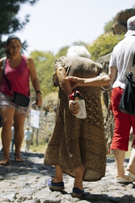 An elderly woman with white hair is walking uphill on a cobblestone path, holding a small bag containing red flowers behind her back. She is wearing a brown patterned dress and blue slippers. On her right, a woman in a pink top with a black bag is walking alongside her, and to the right, a person in a white hat and red shorts is also present. The background shows a sunlit natural environment with trees and rocks.