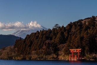 A scenic landscape features a red torii gate standing by a tranquil lake, with a lush forest in the background. The iconic Mount Fuji, capped with snow and partially shrouded by clouds, towers majestically in the distance under a clear blue sky.
