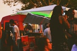 A vibrant food truck scene with people gathered around. The truck is red with a serving hatch open, displaying various food items. Patrons are casually dressed, some with distinctive hairstyles, enjoying a relaxed, social atmosphere under the trees.