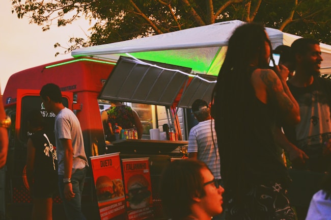 A vibrant food truck scene with people gathered around. The truck is red with a serving hatch open, displaying various food items. Patrons are casually dressed, some with distinctive hairstyles, enjoying a relaxed, social atmosphere under the trees.