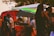 A vibrant food truck scene with people gathered around. The truck is red with a serving hatch open, displaying various food items. Patrons are casually dressed, some with distinctive hairstyles, enjoying a relaxed, social atmosphere under the trees.
