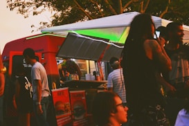 A vibrant food truck scene with people gathered around. The truck is red with a serving hatch open, displaying various food items. Patrons are casually dressed, some with distinctive hairstyles, enjoying a relaxed, social atmosphere under the trees.
