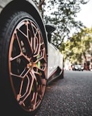 A close-up view of a sports car's bronze alloy wheel with intricate design details, set against an urban street background with trees and another car in the distance.
