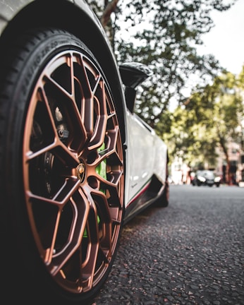 A close-up view of a sports car's bronze alloy wheel with intricate design details, set against an urban street background with trees and another car in the distance.