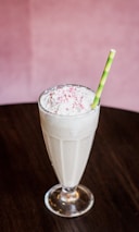 A tall glass filled with a creamy milkshake topped with whipped cream and pink sprinkles. A green-striped straw stands upright in the glass. The milkshake is placed on a dark wooden table, with a soft pink background.