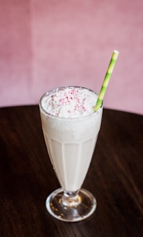A tall glass filled with a creamy milkshake topped with whipped cream and pink sprinkles. A green-striped straw stands upright in the glass. The milkshake is placed on a dark wooden table, with a soft pink background.