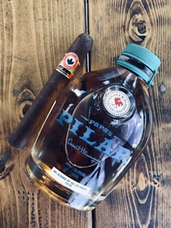 A bottle of Papa's Pilar rum with Ernest Hemingway's signature on it lies on a wooden surface next to a cigar labeled 'Joya de Nicaragua' with a decorative band. The bottle has a teal cap and displays the text 'Blonde Rum' and alcohol content of 46%.