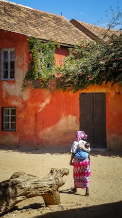 woman with baby on back walking on pathway near orange concrete house under blue and white skies during daytime