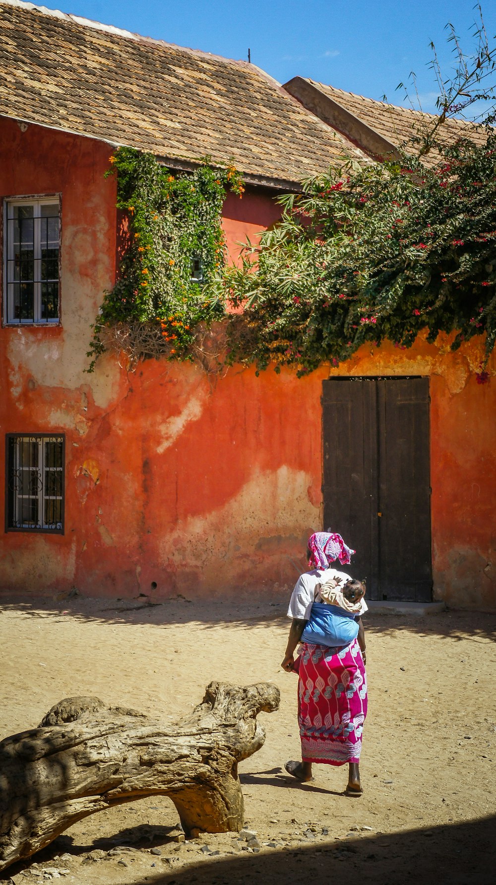 昼間、青と白の空の下、オレンジ色のコンクリートの家の近くの小道を歩く背中に赤ん坊を乗せた女性