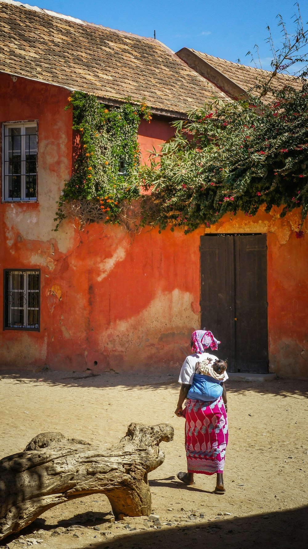 woman with baby on back walking on pathway near orange concrete house under blue and white skies during daytime