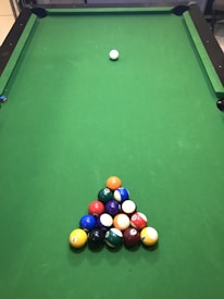 A green pool table with a neatly arranged triangular rack of colorful billiard balls positioned near one end. A single cue ball is placed further up the table, away from the rack.