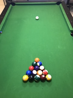 A green pool table with a neatly arranged triangular rack of colorful billiard balls positioned near one end. A single cue ball is placed further up the table, away from the rack.