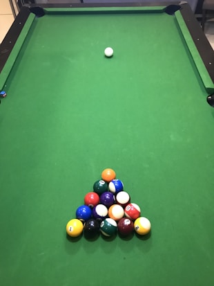 A green pool table with a neatly arranged triangular rack of colorful billiard balls positioned near one end. A single cue ball is placed further up the table, away from the rack.