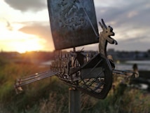 A metal sculpture of a Viking ship with a dragon-shaped prow is set against a sunset backdrop. The ship features intricate details, including shields along the sides and extended oars. The landscape in the distance is partially blurred, adding focus to the sculpture.