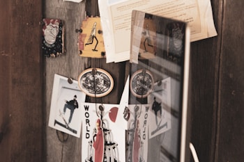 A collection of eclectic images and memorabilia pinned on a wooden board. Among them are photographs of a skateboarder, a piece of art with the word 'WORLD', and various small pieces of paper or stickers. There is a reflection of these items on a nearby reflective surface, possibly a glass or mirror, enhancing the visual depth.