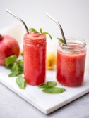 Two glass jars filled with a vibrant red smoothie are placed on a white marble surface. Each jar has a metal straw and is garnished with fresh mint leaves. In the background, a pomegranate, a lemon, and additional mint leaves are visible, adding a fresh and natural feel to the setting.