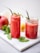 Two glass jars filled with a vibrant red smoothie are placed on a white marble surface. Each jar has a metal straw and is garnished with fresh mint leaves. In the background, a pomegranate, a lemon, and additional mint leaves are visible, adding a fresh and natural feel to the setting.