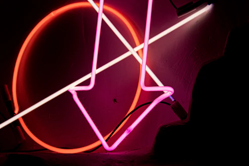 Close-up of a designer sketching neon sign concepts on a dark desk illuminated by colorful neon lights.