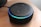 A black smart speaker device resting on a wooden surface, featuring a glowing blue light ring on top with control buttons visible, including plus, minus, and other symbols.