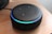 A black smart speaker device resting on a wooden surface, featuring a glowing blue light ring on top with control buttons visible, including plus, minus, and other symbols.