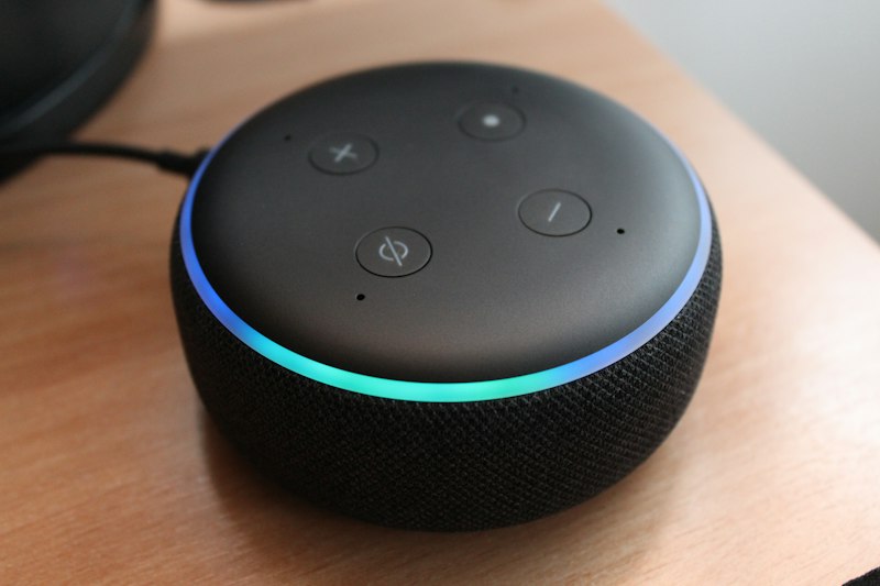 An Amazon Echo Dot smart speaker sitting on a wooden desk beside a computer mouse