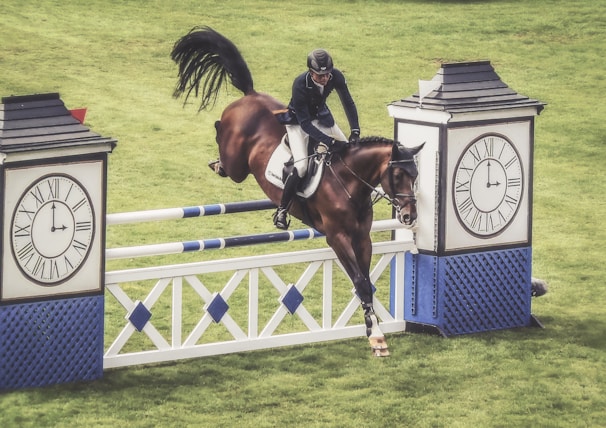 A horse and rider are captured mid-jump over a show jumping obstacle designed to look like two large clocks. The grassy field sets a natural backdrop, and the rider is dressed in typical equestrian attire including a helmet. The brown horse is equipped with a saddle and bridle.
