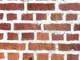 A close-up view of a brick wall with red bricks and gray mortar. The bricks are arranged in a staggered pattern typical of traditional masonry work.