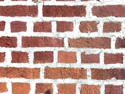 A close-up view of a brick wall with red bricks and gray mortar. The bricks are arranged in a staggered pattern typical of traditional masonry work.