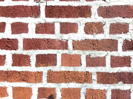 A close-up view of a brick wall with red bricks and gray mortar. The bricks are arranged in a staggered pattern typical of traditional masonry work.