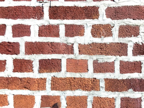 A close-up view of a brick wall with red bricks and gray mortar. The bricks are arranged in a staggered pattern typical of traditional masonry work.