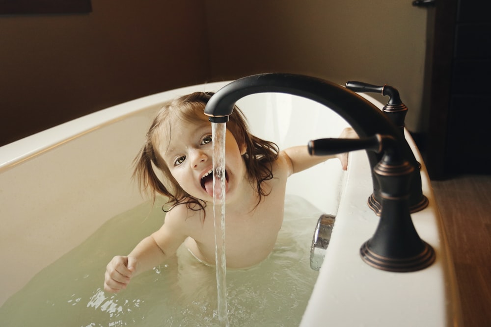 girl in bathtub tasting water from black faucet
