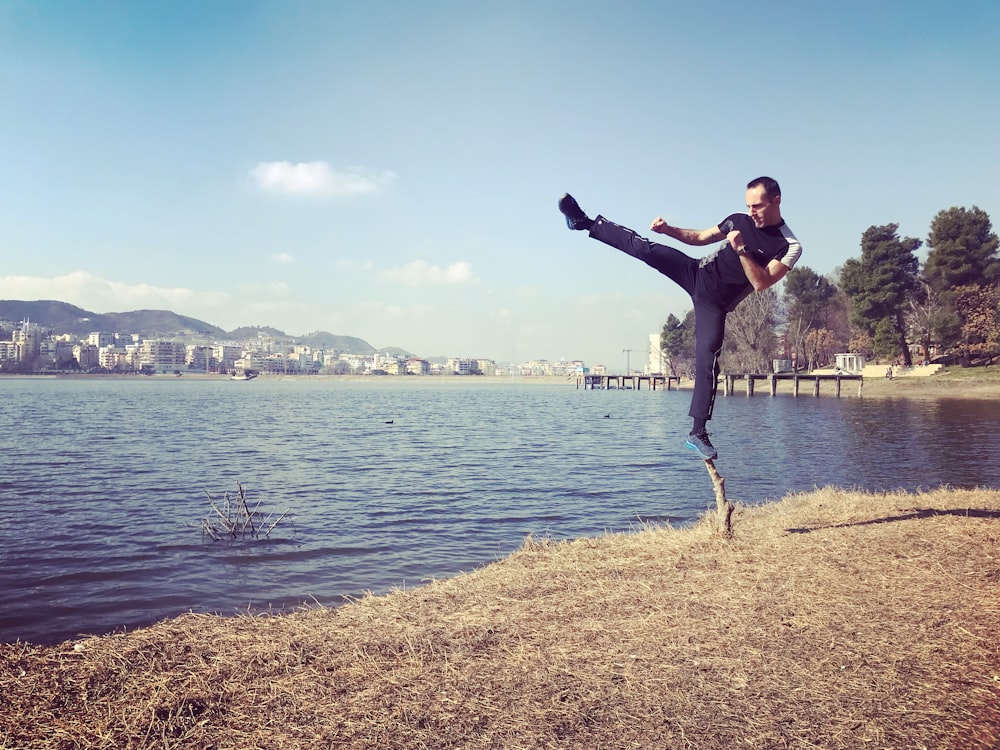 hombre de pie sobre la pierna en la ramita haciendo la pose de patada al lado del cuerpo de agua durante el día