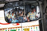 A bus filled with passengers is captured in a rearview mirror, where a man in a checkered shirt is driving. The passengers are seated, some looking out the windows, with a variety of expressions. There are colorful decorations hanging in front of the mirror.