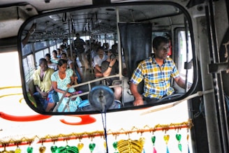 A bus filled with passengers is captured in a rearview mirror, where a man in a checkered shirt is driving. The passengers are seated, some looking out the windows, with a variety of expressions. There are colorful decorations hanging in front of the mirror.
