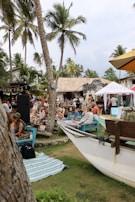 A lively outdoor gathering featuring numerous people seated on wooden benches under tall palm trees. The scene is set in a tropical locale with a mix of rustic buildings and modern elements. Colorful blankets and beach towels are spread on the grass, and a small boat is positioned in the foreground.